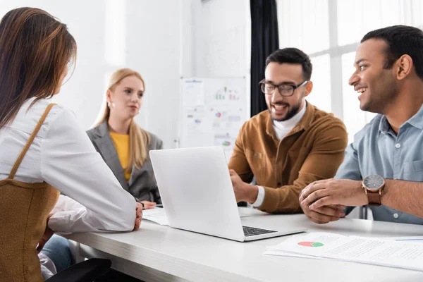 Laptop near cheerful multiethnic businesspeople on blurred background — Stock Photo