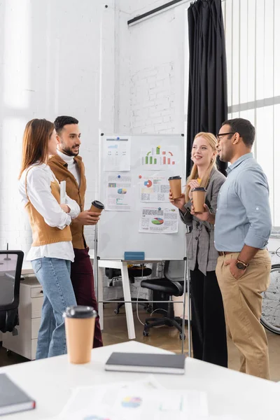 Cheerful multiethnic businesspeople with takeaway drink working near charts on flipchart — Stock Photo