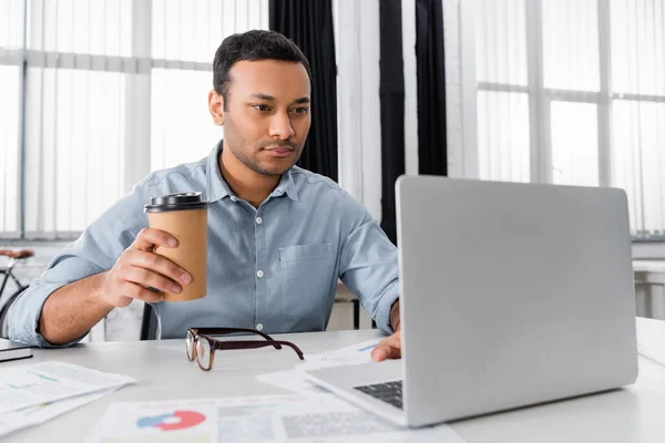 Indian businessman with takeaway drink using laptop on blurred foreground in office — Stock Photo