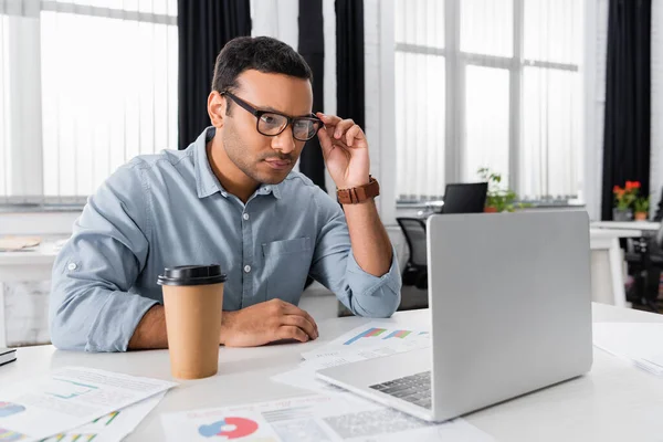 Indischer Geschäftsmann blickt auf Laptop in der Nähe von Coffee to go und Papiere im verschwommenen Vordergrund — Stockfoto