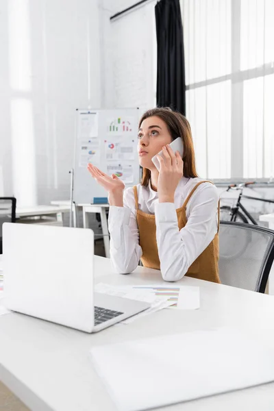 Empresária falando no smartphone perto do laptop em primeiro plano desfocado — Fotografia de Stock