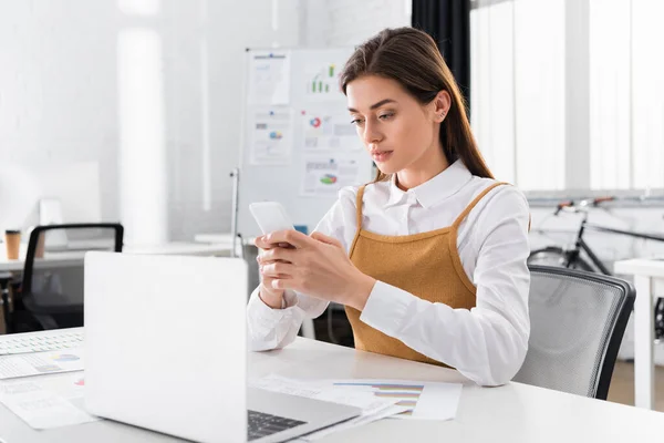 Junge Geschäftsfrau mit Smartphone in der Nähe von Dokumenten und Laptop im verschwommenen Vordergrund — Stockfoto