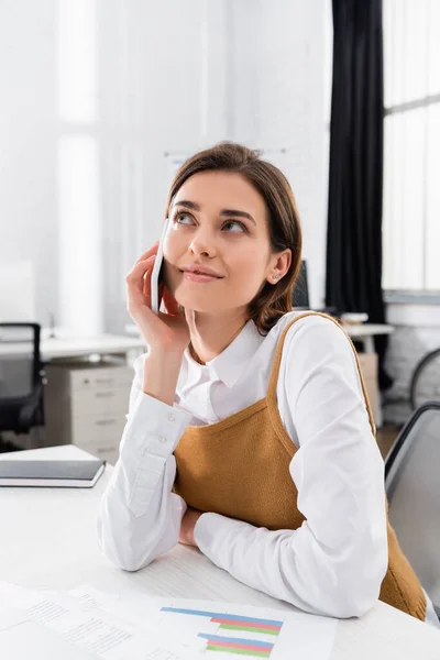 Lächelnde Geschäftsfrau spricht auf Smartphone neben Zeitungen mit Diagrammen im verschwommenen Vordergrund — Stockfoto