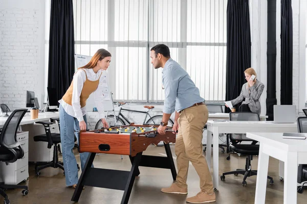 Interracial business people playing table soccer near colleague talking on smartphone in office — Stock Photo