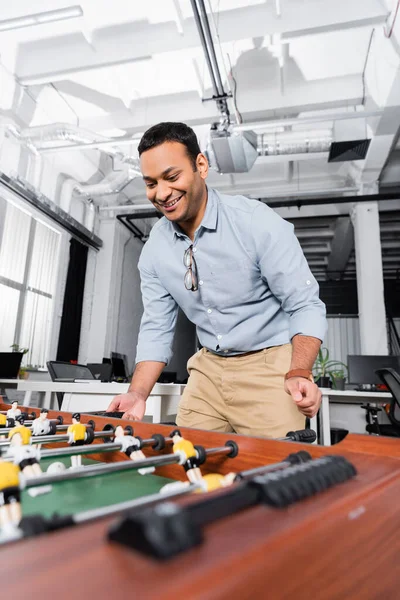 Cheerful indian businessman playing table soccer on blurred foreground in office — Stock Photo