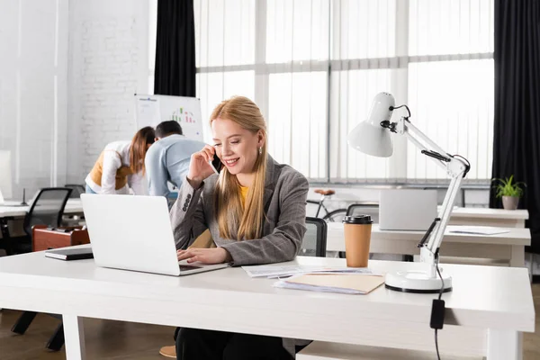 Femme d'affaires souriante parlant sur smartphone près d'un ordinateur portable et café pour aller au bureau — Photo de stock