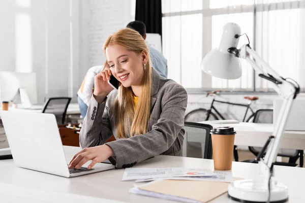 Donna d'affari sorridente che usa il computer portatile e parla su smartphone vicino a documenti e lampada in ufficio — Foto stock