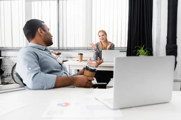 Mujer de negocios sonriente hablando con colega indio con café para ir cerca de la computadora portátil en la oficina - foto de stock