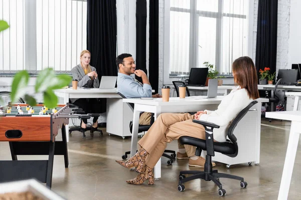 Hombre de negocios indio sonriente sentado cerca de colegas y café para ir en la oficina - foto de stock