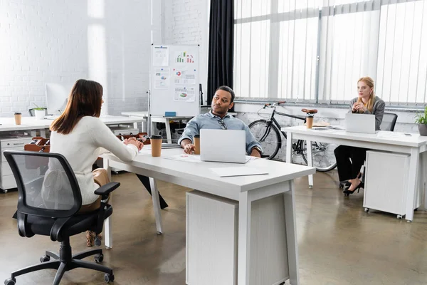 Empresarios multiétnicos hablando cerca del café para ir y portátiles en la oficina - foto de stock