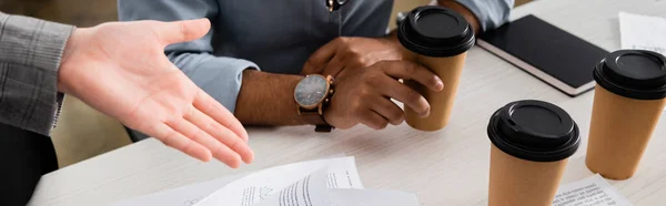 Vue recadrée d'une femme d'affaires pointant vers des papiers près d'un collègue indien avec du café à emporter, bannière — Photo de stock