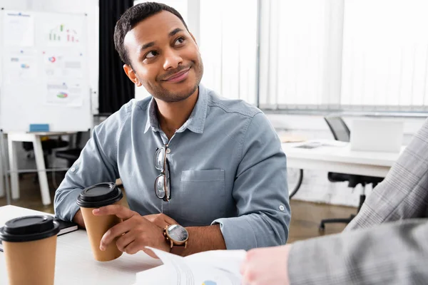 Lächelnder indischer Geschäftsmann mit Kaffee in der Hand nähert sich Kollegen auf verschwommenem Vordergrund — Stockfoto