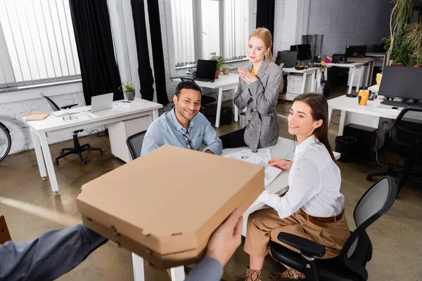 Pessoas de negócios multiétnicos sorridentes olhando para o homem de entrega com caixas de pizza em primeiro plano borrado — Fotografia de Stock