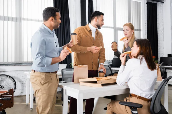 Мультиэтнические бизнесмены едят пиццу и держат кофе во время обеда — стоковое фото