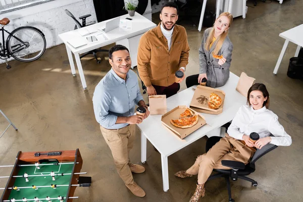 Vista aérea de gente de negocios multiétnicos sonriendo cerca de pizza y café para ir en la oficina - foto de stock