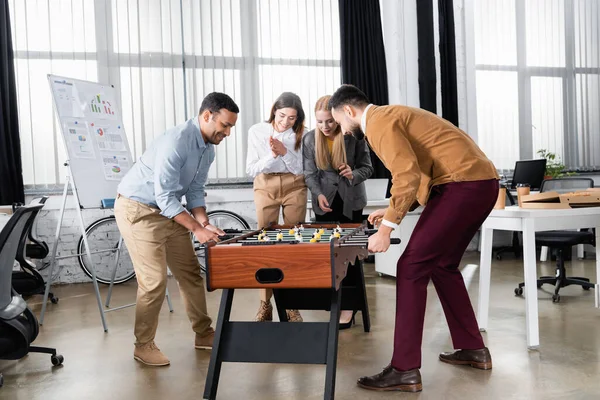 Веселые многонациональные бизнесмены играют в настольный футбол рядом с улыбающимися деловыми женщинами в офисе — стоковое фото