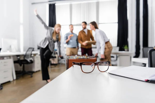 Eyeglasses and notebook on table near business people on blurred background in office — Stock Photo