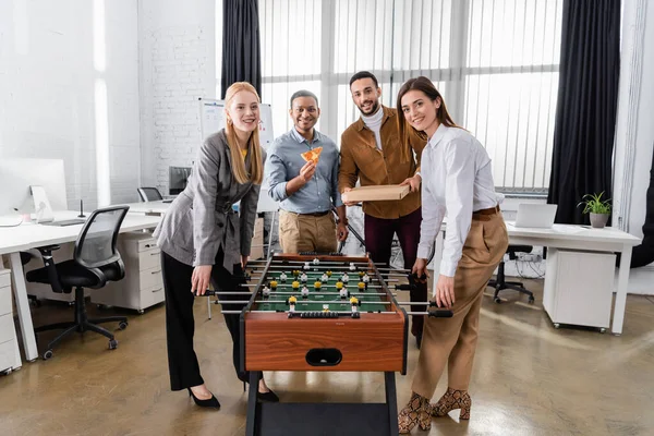 Des hommes d'affaires interraciaux souriants avec une pizza regardant une caméra près du baby-foot au bureau — Photo de stock