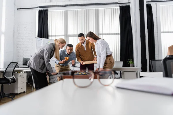 Multiethnic business people with pizza playing table soccer near eyeglasses on blurred foreground in office — Stock Photo