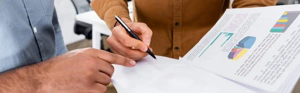 Vista recortada del hombre de negocios señalando con el dedo cerca de colega con papeles y pluma en la oficina, pancarta — Stock Photo