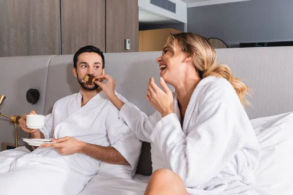 Cheerful woman feeding croissant to muslim boyfriend with coffee on hotel bed — Stock Photo