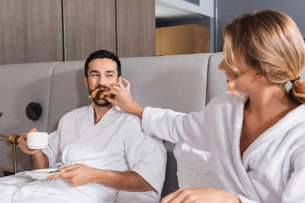 Jeune femme nourrissant croissant au petit ami arabe avec tasse sur le lit à l'hôtel — Photo de stock