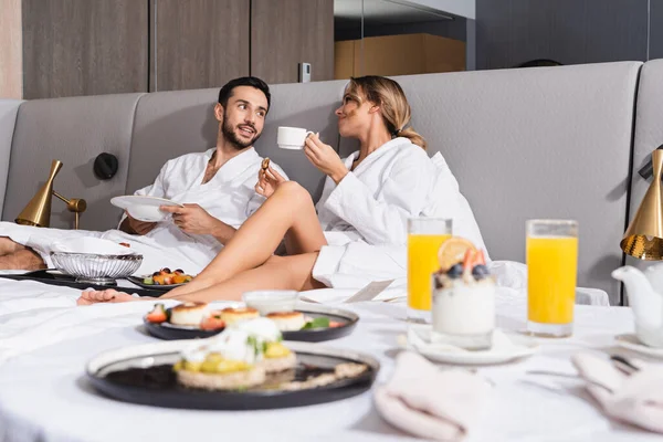 Smiling interracial couple looking at each other near delicious breakfast on blurred foreground on hotel bed — Stock Photo