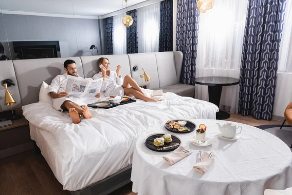 Muslim man reading newspaper near girlfriend talking on smartphone and delicious breakfast in hotel room — Stock Photo