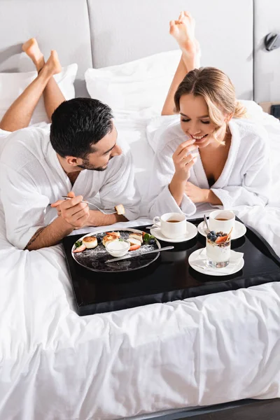 Mulher sorridente olhando para sobremesas na bandeja perto de namorado muçulmano na cama do hotel — Fotografia de Stock