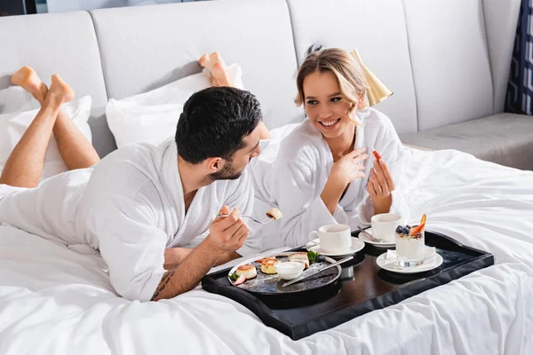 Cheerful woman in bathrobe looking at muslim boyfriend with fork near delicious breakfast on hotel bed — Stock Photo
