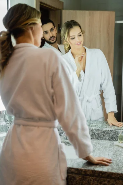 Mulher sorridente em roupão de banho perto do espelho e namorado muçulmano no banheiro do hotel — Fotografia de Stock