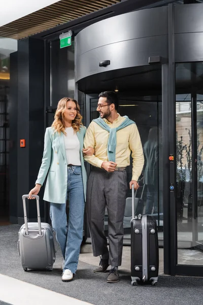 Mujer positiva caminando cerca de novio musulmán con maleta cerca del hotel — Stock Photo