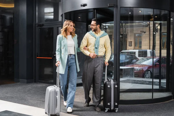 Junges gemischtrassiges Paar lächelt einander an, während es mit Gepäck in Hotelnähe spaziert — Stockfoto