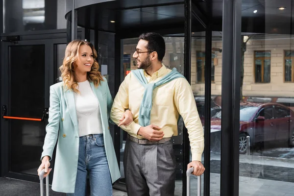 Cheerful woman smiling at muslim boyfriend with suitcase near hotel — Stock Photo