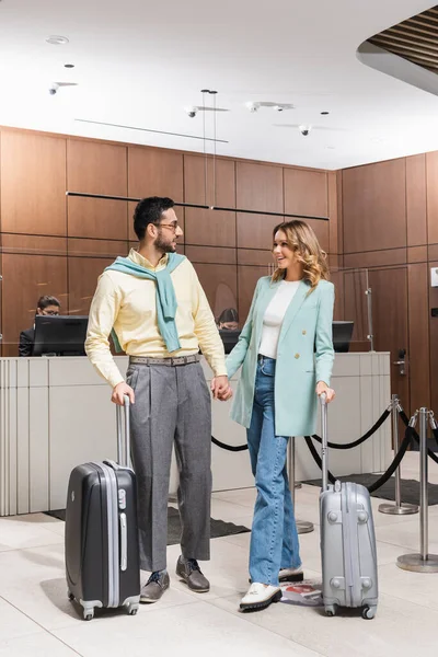 Smiling woman with suitcase looking at muslim boyfriend during vacation in hotel — Stock Photo