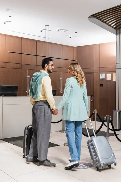 Smiling woman holding hand of muslim boyfriend with suitcase in hotel — Stock Photo