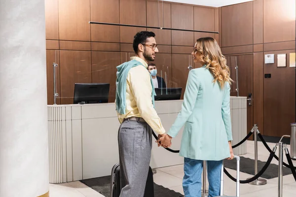Couple interracial souriant de touristes tenant la main dans le hall de l'hôtel — Photo de stock