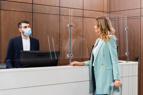 Receptionist in maschera medica guardando la donna con valigia nella hall dell'hotel — Foto stock