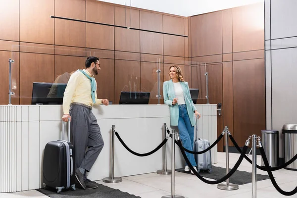 Smiling interracial couple with suitcases waiting near hotel reception — Stock Photo