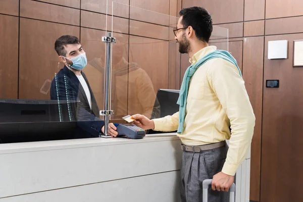 Muslim man paying with credit card near manager in medical mask with payment terminal in hotel — Stock Photo