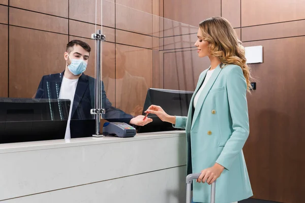 Femme souriante avec clé de prise de bagages du directeur de l'hôtel dans le masque médical dans le hall — Photo de stock