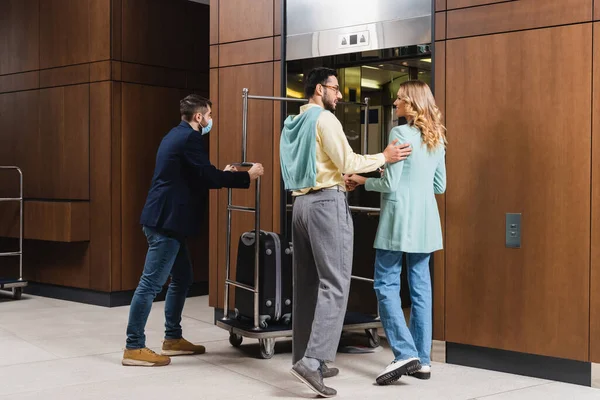 Interracial couple standing near hotel porter in medical mask and elevator — Stock Photo