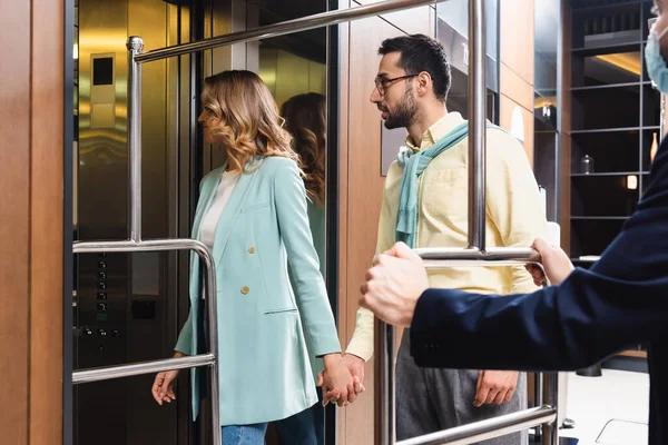 Casal inter-racial em pé perto do elevador e porteiro em máscara médica em primeiro plano desfocado no hotel — Fotografia de Stock