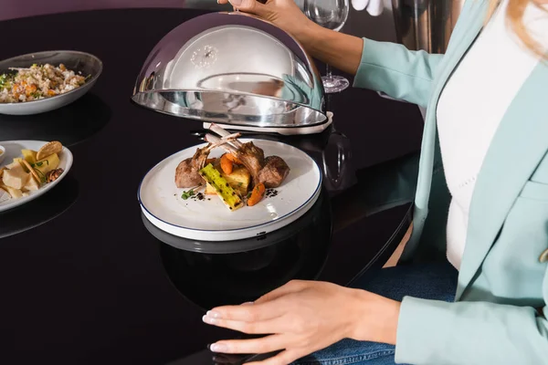 Vista recortada de la mujer sosteniendo cloche cerca de delicioso plato en la habitación del hotel - foto de stock