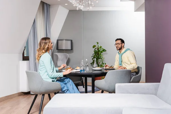 Sonriente pareja interracial hablando durante la cena en la habitación del hotel - foto de stock