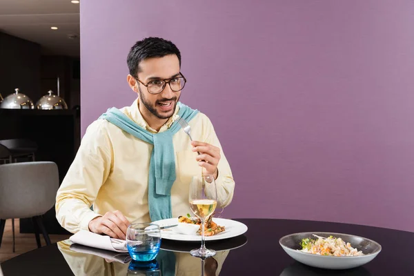 Hombre árabe sonriente sosteniendo tenedor cerca de la comida y el vino en la habitación del hotel - foto de stock