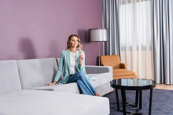 Mujer sonriente con copa de vino sentada en un sofá en una habitación de hotel moderna - foto de stock