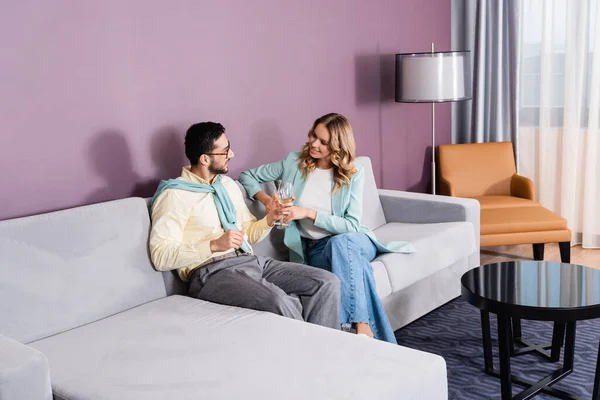 Smiling interracial couple holding glasses of wine on couch in hotel — Stock Photo