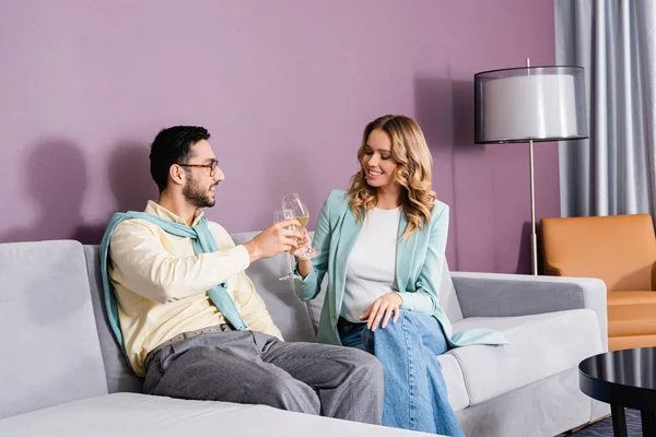 Pareja interracial brindando con vino en habitación de hotel - foto de stock