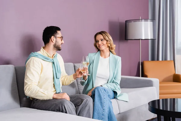 Sonriente pareja interracial con copas de vino sentado en el sofá en la habitación de hotel - foto de stock